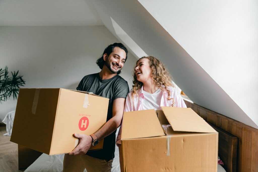 A smiling couple holding cardboard moving boxes in a room with slanted white walls.