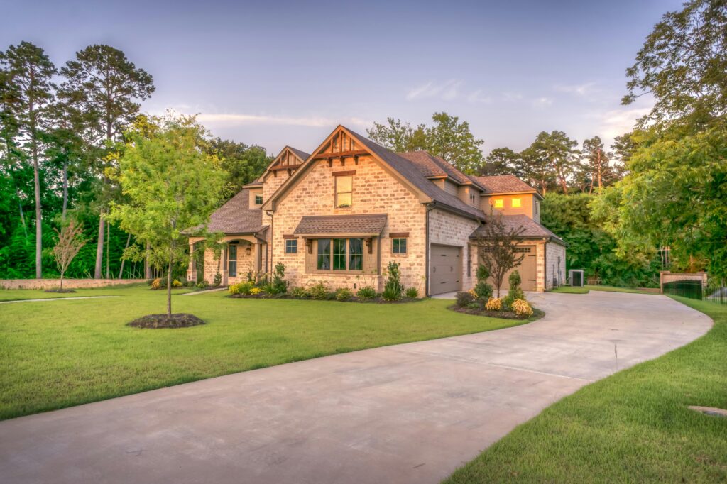 A stone and brick two-story house with a steep gabled roof, arched entryway, and attached garage, surrounded by a manicured green lawn and trees at dusk.