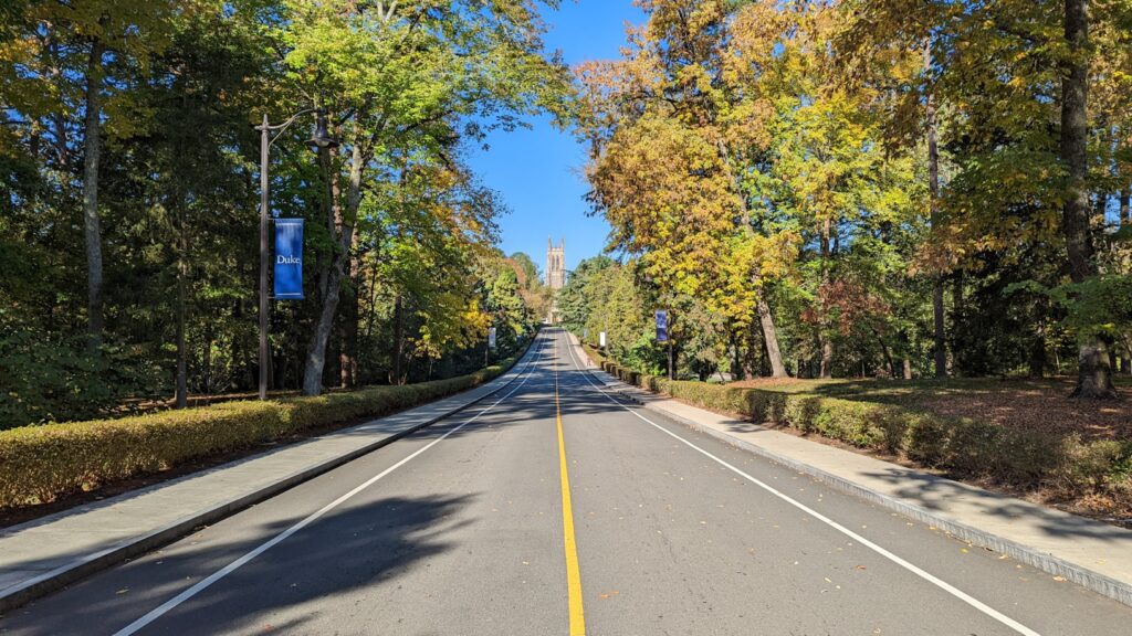 A tree-lined road in autumn with yellow and orange foliage leading toward Duke University's campus, with a blue banner on a light pole and the Duke Chapel tower visible in the distance.
