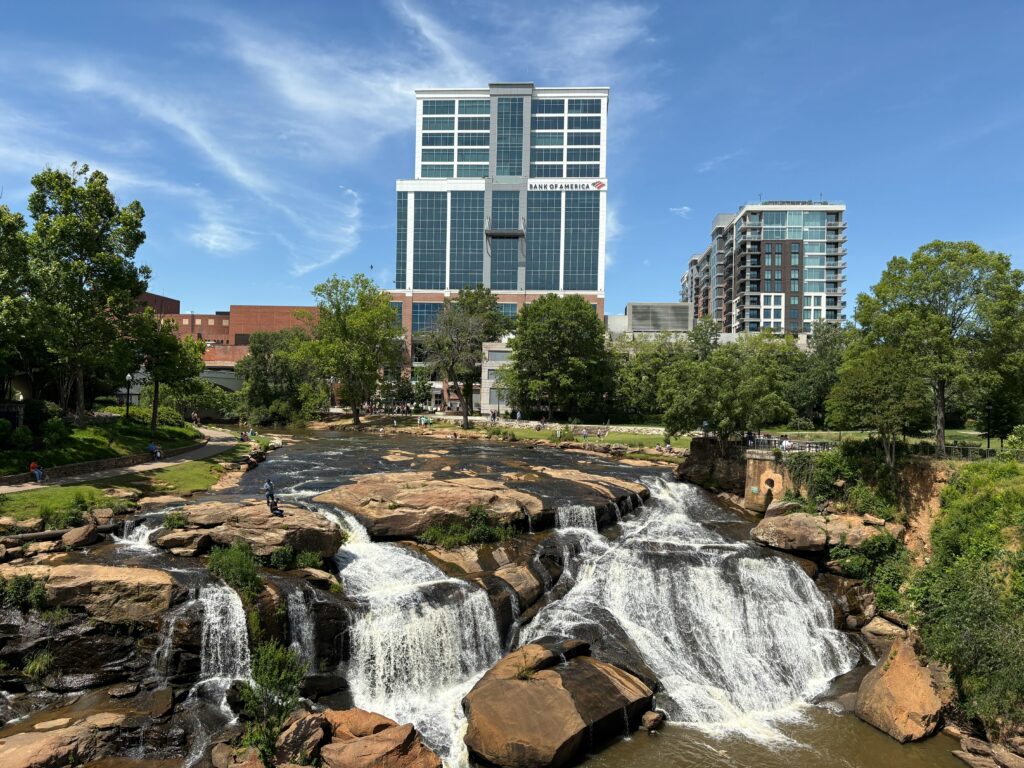 Falls Park on the Reedy in Greenville, South Carolina, with a cascading waterfall flowing over rocks in front of modern high-rise buildings and green trees.