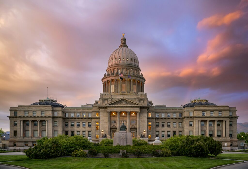 A grand state capitol building with a central dome and symmetrical wings, lit warmly against a purple and pink cloudy sky at dusk.