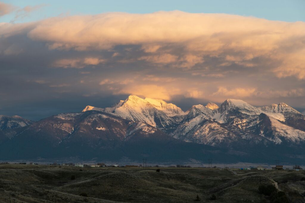 Snow-capped mountain peaks with golden sunset light breaking through heavy clouds above brown rolling foothills.