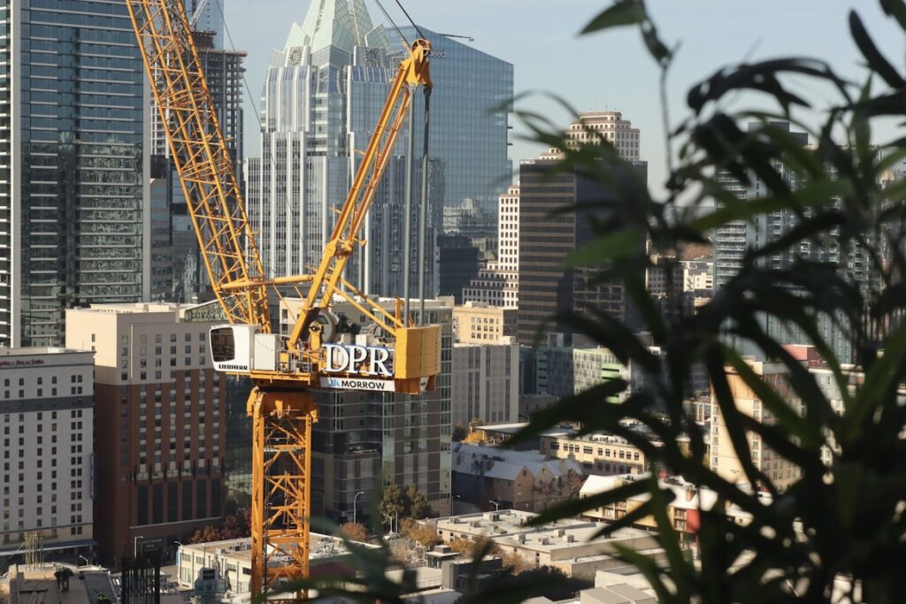 A yellow DPR Construction crane in the foreground with the Austin, Texas skyline and high-rise buildings behind it on a hazy day, with a plant partially framing the right side.