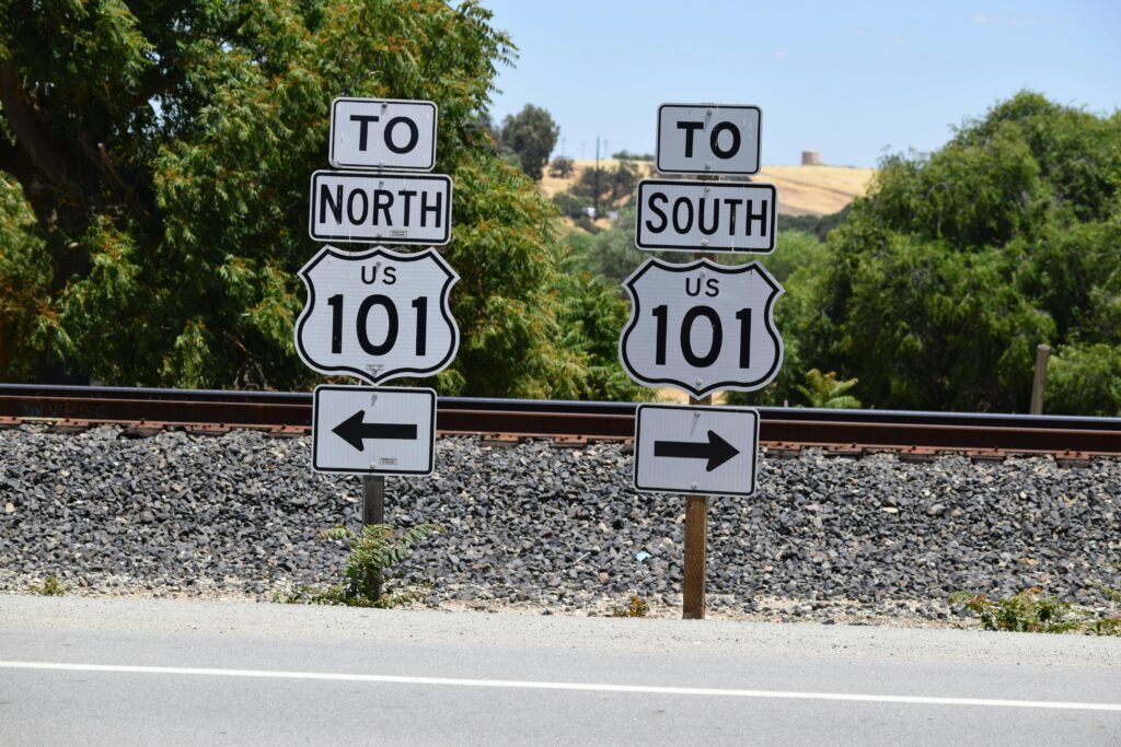 Two US Route 101 highway signs on metal posts, one pointing left to North and the other pointing right to South, with railroad tracks and dry golden hills in the background.