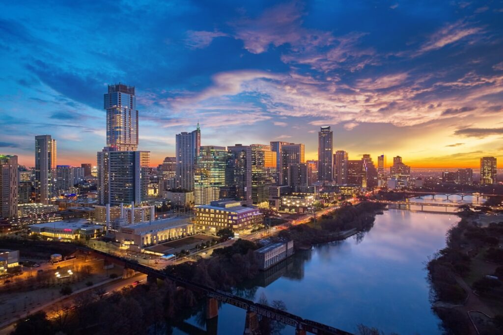 Austin, Texas skyline at sunset with high-rise buildings reflecting orange and blue light, overlooking the Colorado River and a railroad bridge.