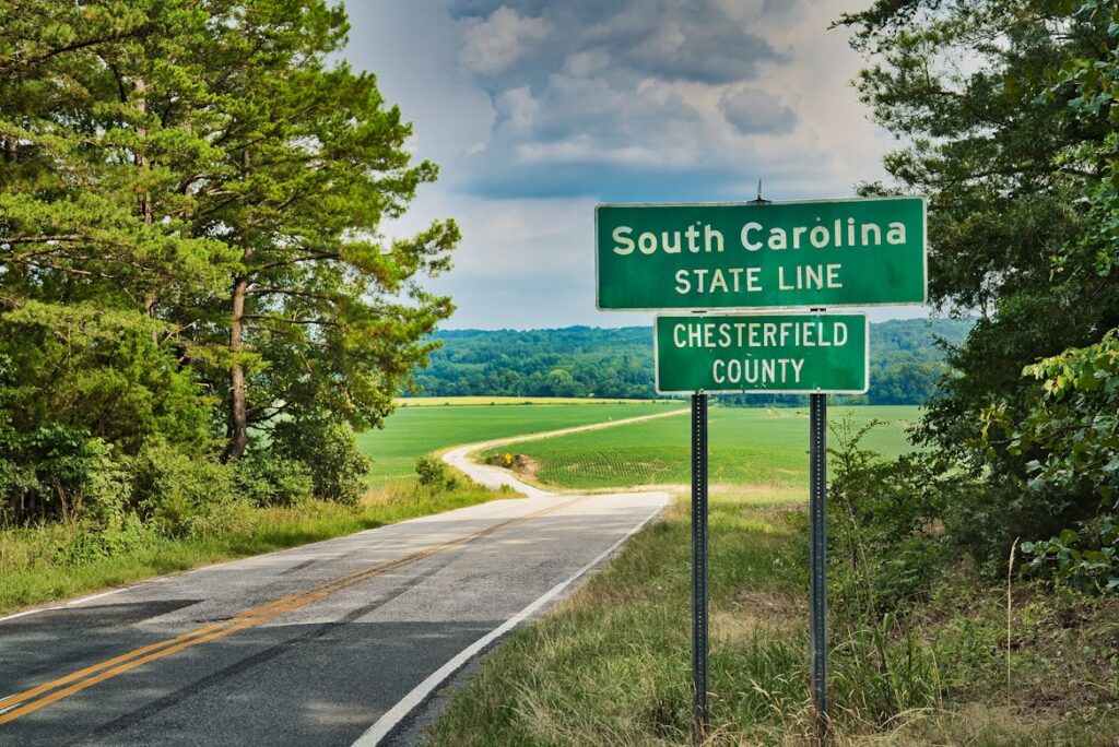 A green "South Carolina State Line, Chesterfield County" highway sign on a two-lane road lined with trees and green fields.