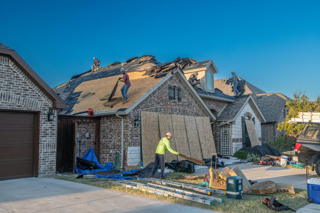 Roofing workers replacing shingles on a gray brick suburban home, with torn-off materials and blue tarps on the ground below.