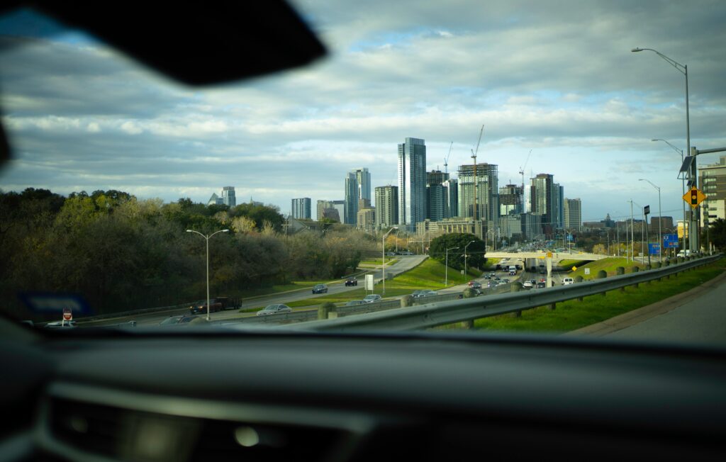 A city skyline with construction cranes viewed through a car windshield while driving on a highway on an overcast day.