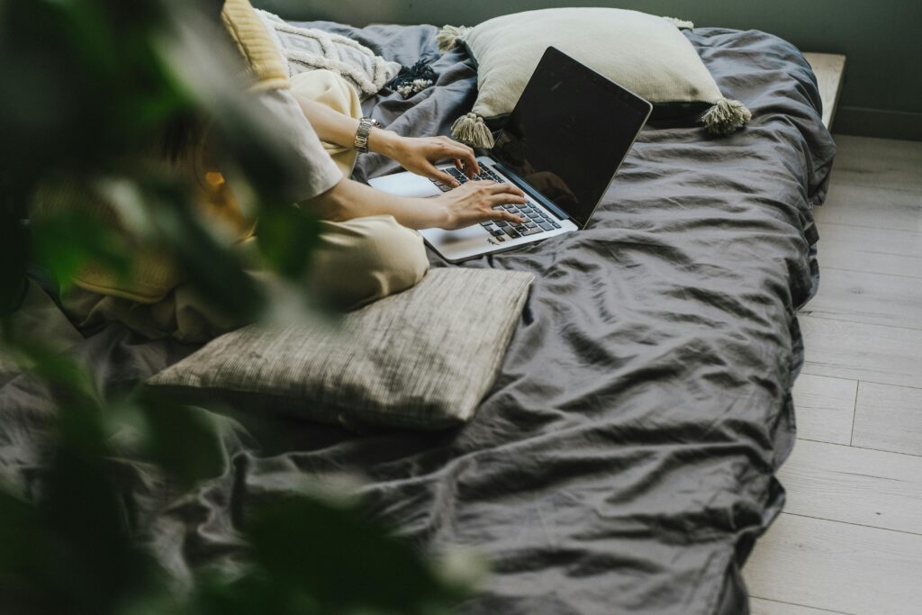 A person sitting on a bed with gray bedding, typing on a laptop, partially obscured by an indoor plant in the foreground.