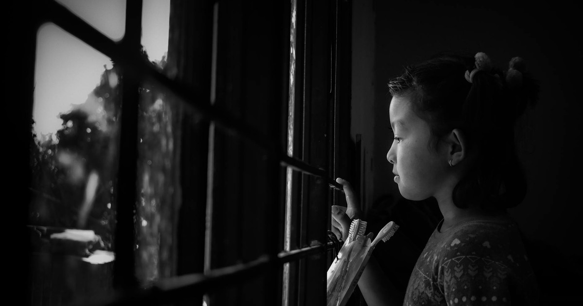 Side view of black and white adorable pensive little Asian girl standing in bathroom and looking out fenced window on rainy day