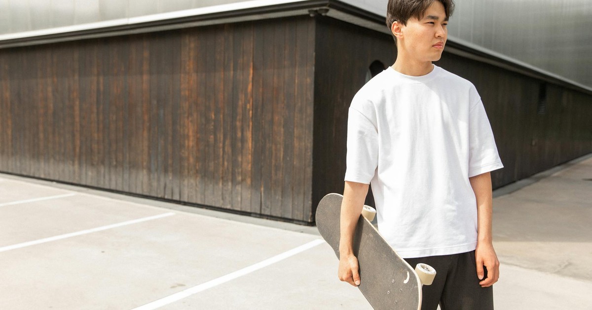 Handsome young ethnic guy in white t shirt holding skateboard in hand and looking away thoughtfully while resting near modern building after ride