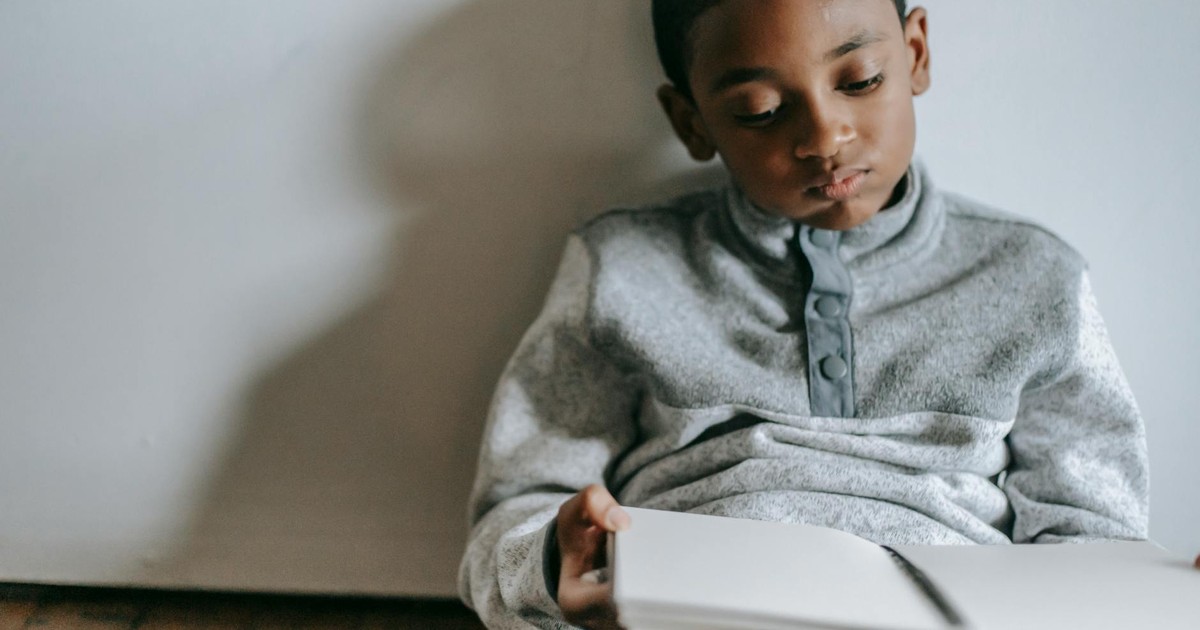 Concentrated African American boy looking at opened copybook while sitting on parquet at home