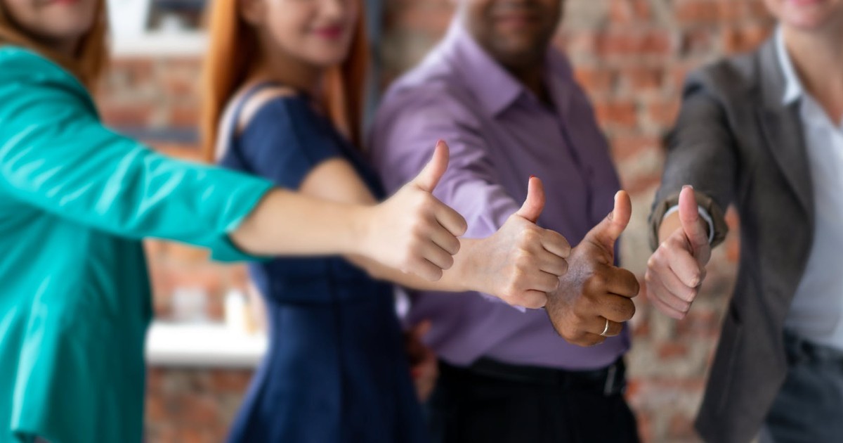 Unrecognizable cheerful colleagues in formal clothes showing thumbs up gesture while standing in modern office during teamwork on blurred background