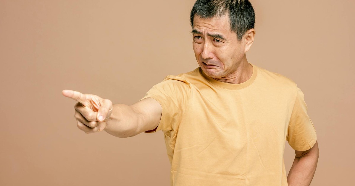 An adult man with a frustrated expression points his finger in anger against a brown background.