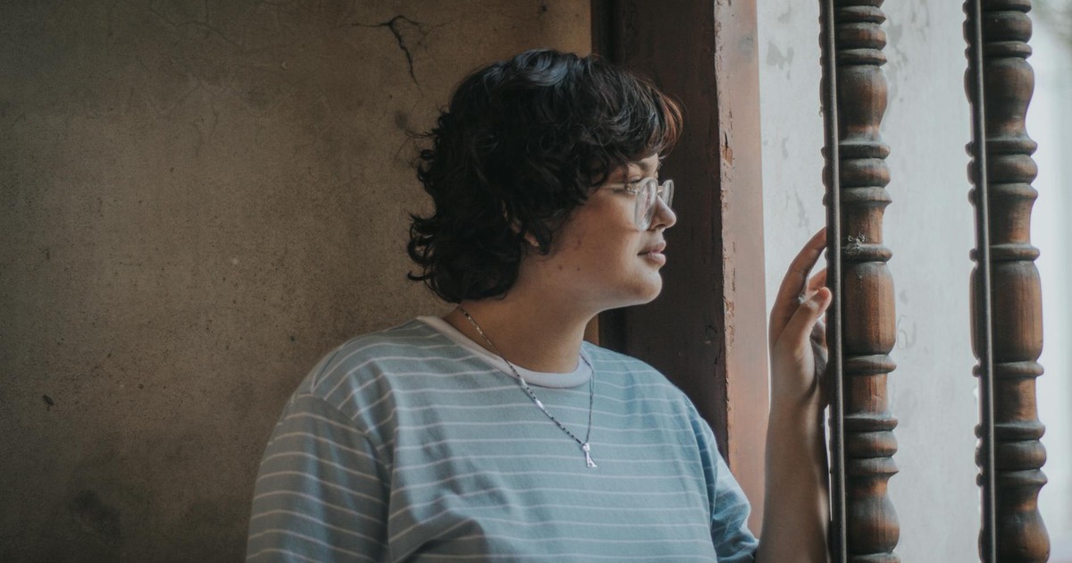 A thoughtful woman with short hair and eyeglasses gazes out a window.