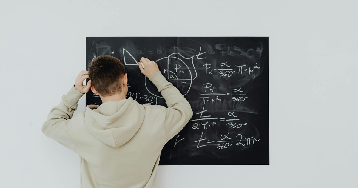Teenager in hoodie writing math formulas on a blackboard indoors, showcasing problem-solving skills.