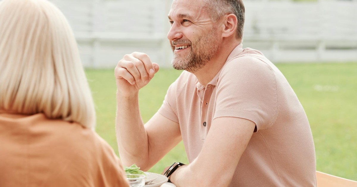 An adult man smiling while seated outdoors, enjoying a casual conversation.