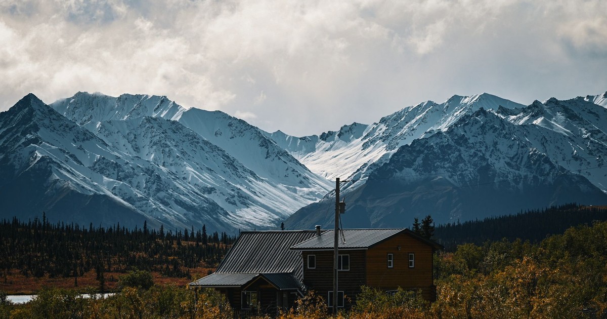 Rustic cabin nestled in lush greenery with majestic snow-capped mountains.