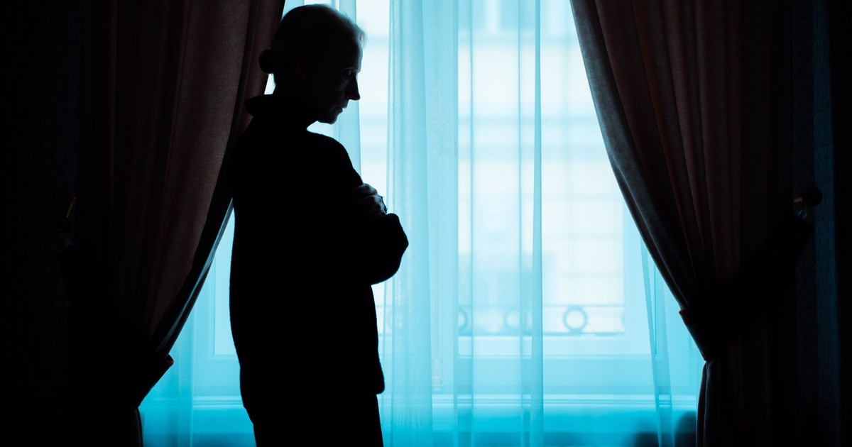 A silhouette of a senior woman stands thoughtfully by a window, conveying solitude.