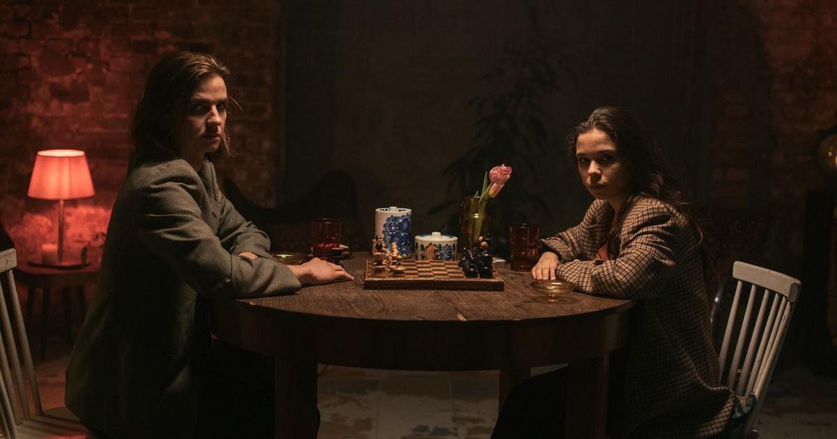 Two women play chess in a dimly lit room with a warm, moody ambiance.