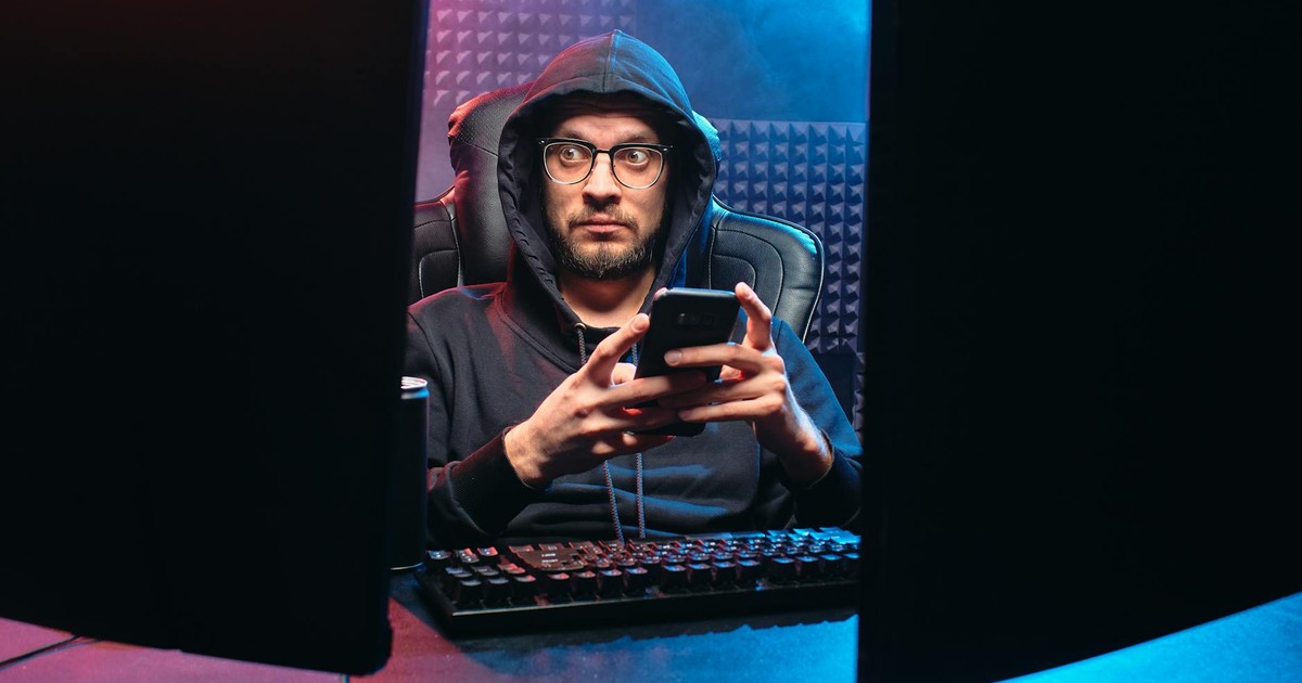 A man in a hoodie using a smartphone, surrounded by tech gear in a dimly lit room.