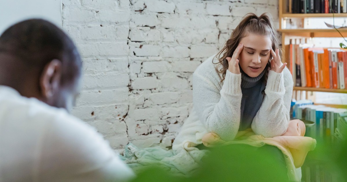 Exhausted woman calming down after argument with husband by putting fingers on temples and man sitting and looking down