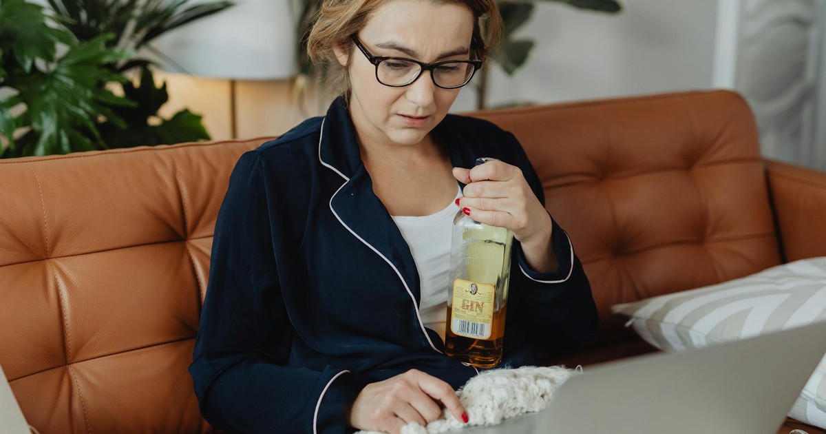 Woman in pajamas holding a bottle of alcohol while working on a laptop, sitting on a sofa at home.