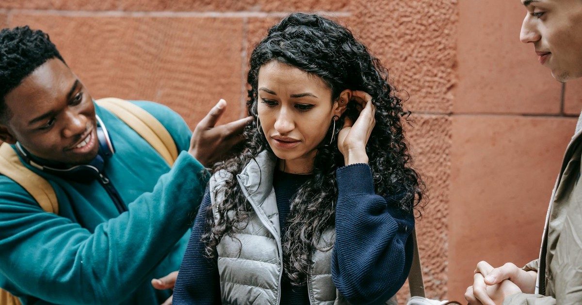 Crop African American teenage man abusing and touching hair of depressed ethnic female groupmate while standing on street with male friend