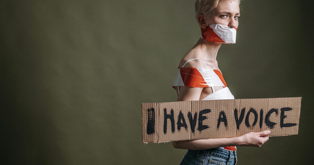 A woman wrapped in tape holds a protest placard saying 'I Have a Voice'.