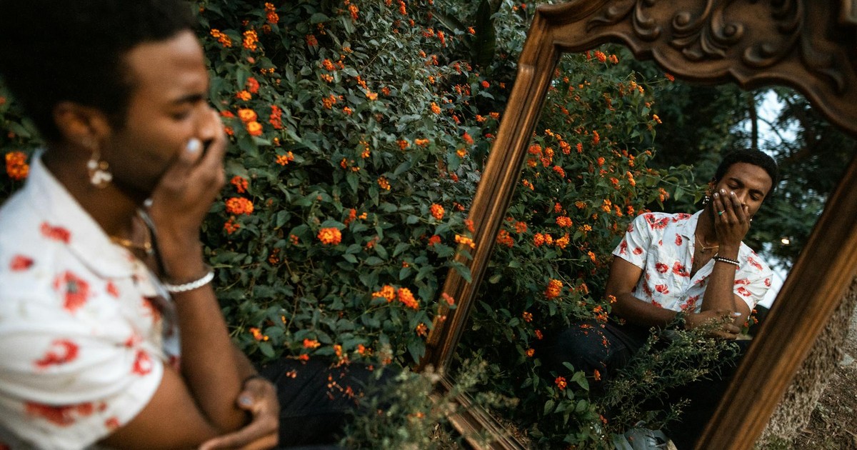 A man in floral shirt sits with a mirror reflecting his contemplative expression amidst orange flowers.