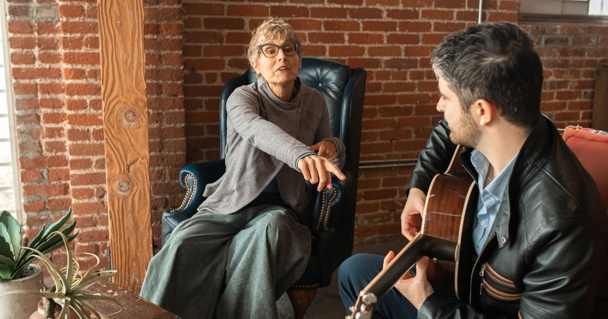 A woman and man share a relaxed moment indoors as he plays guitar and they converse.