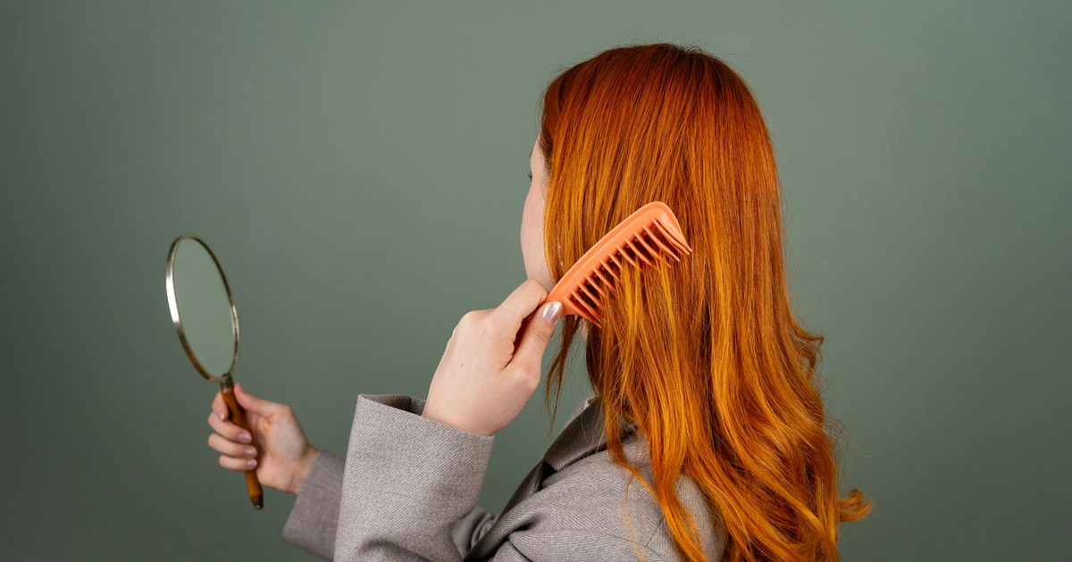 Red-haired woman using a comb while holding a mirror, focusing on hair care and beauty.