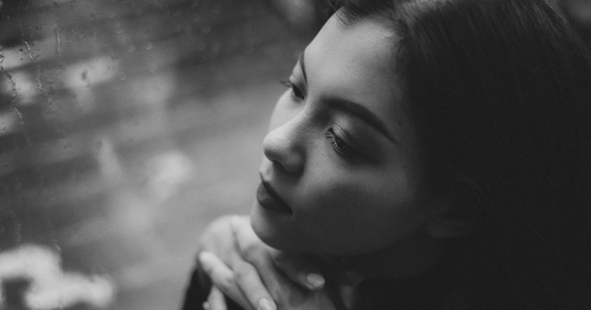 Black and white high angle of serious teenage girl sitting in solitude near window covered with raindrops