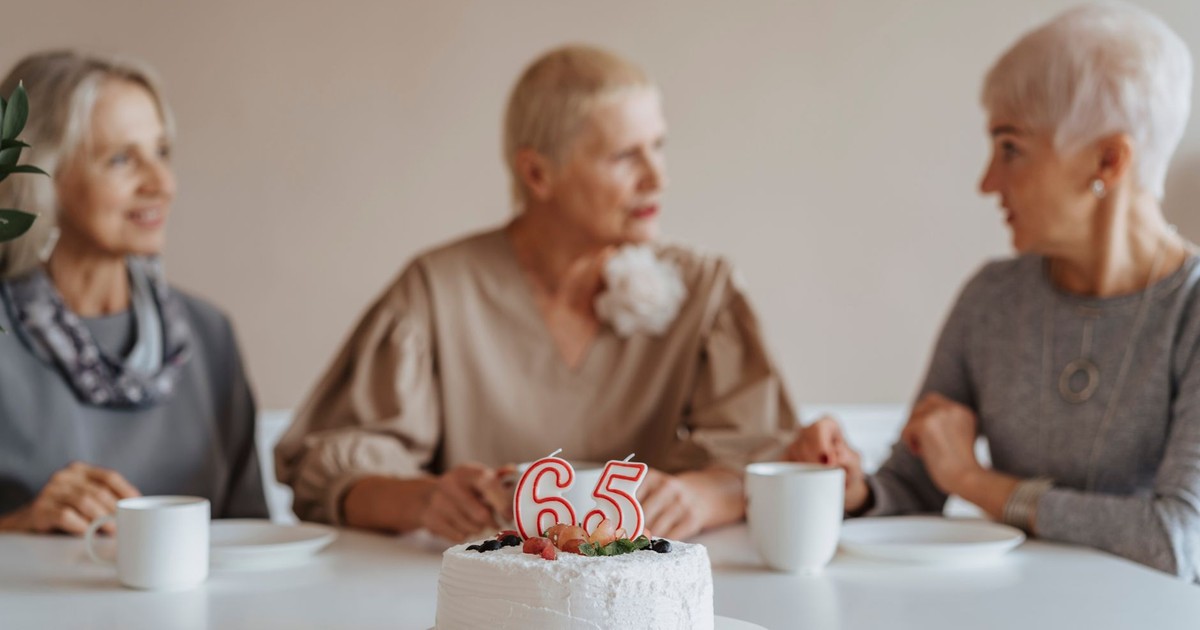 Three senior women celebrate a 65th birthday with cake and coffee indoors.
