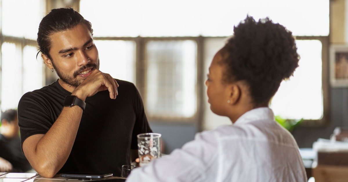 Two adults conversing at a table indoors, with natural light through windows.