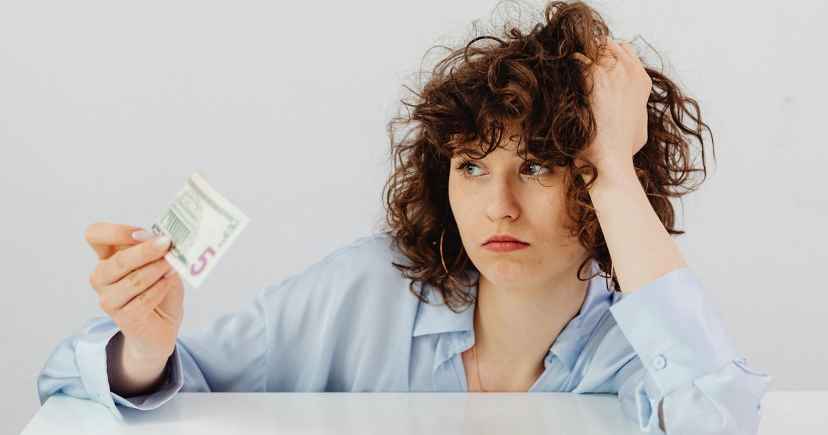 Concerned woman with curly hair holding a five dollar bill, expressing financial worry.