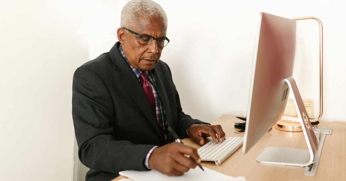 Elderly businessman in suit working at desk with computer and notes in modern office setting.
