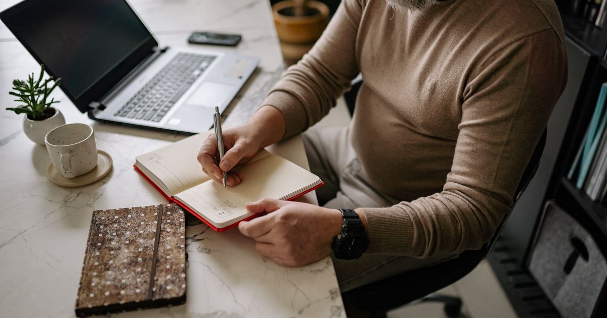 Adult man writing in notebook with laptop and coffee on desk in a cozy indoor setting.