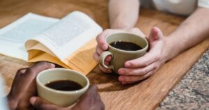 Two people holding coffee cups and reading a book on a wooden table.