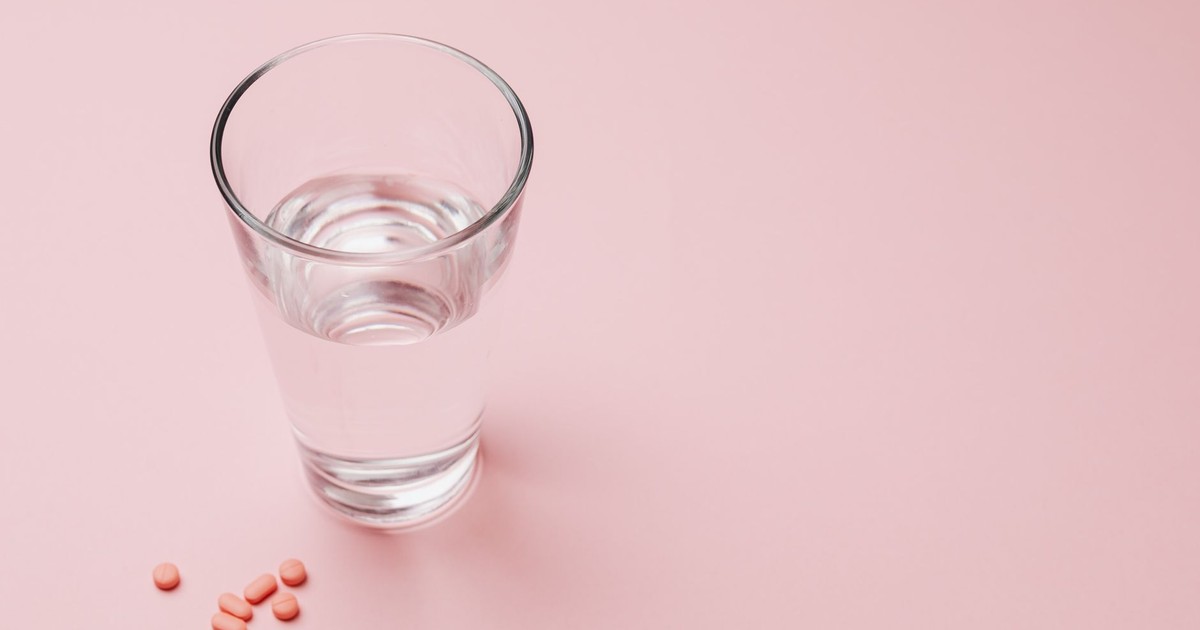 Elevated view of a glass of water next to pink pills on a pastel pink background symbolizing health and wellness.