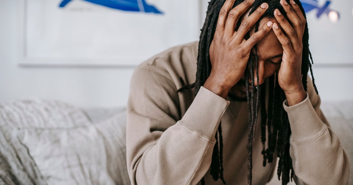 Crop anxious African American male with dreadlocks touching head with closed eyes on couch in house