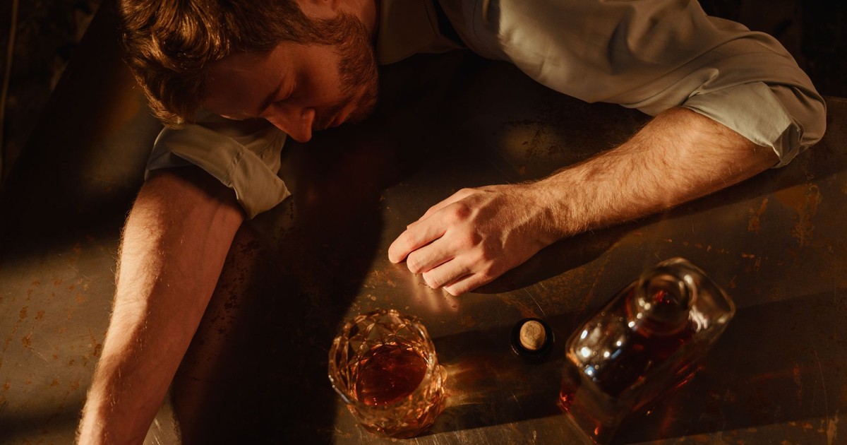 A man sleeps on a table with a whiskey glass and bottle nearby, suggesting exhaustion.