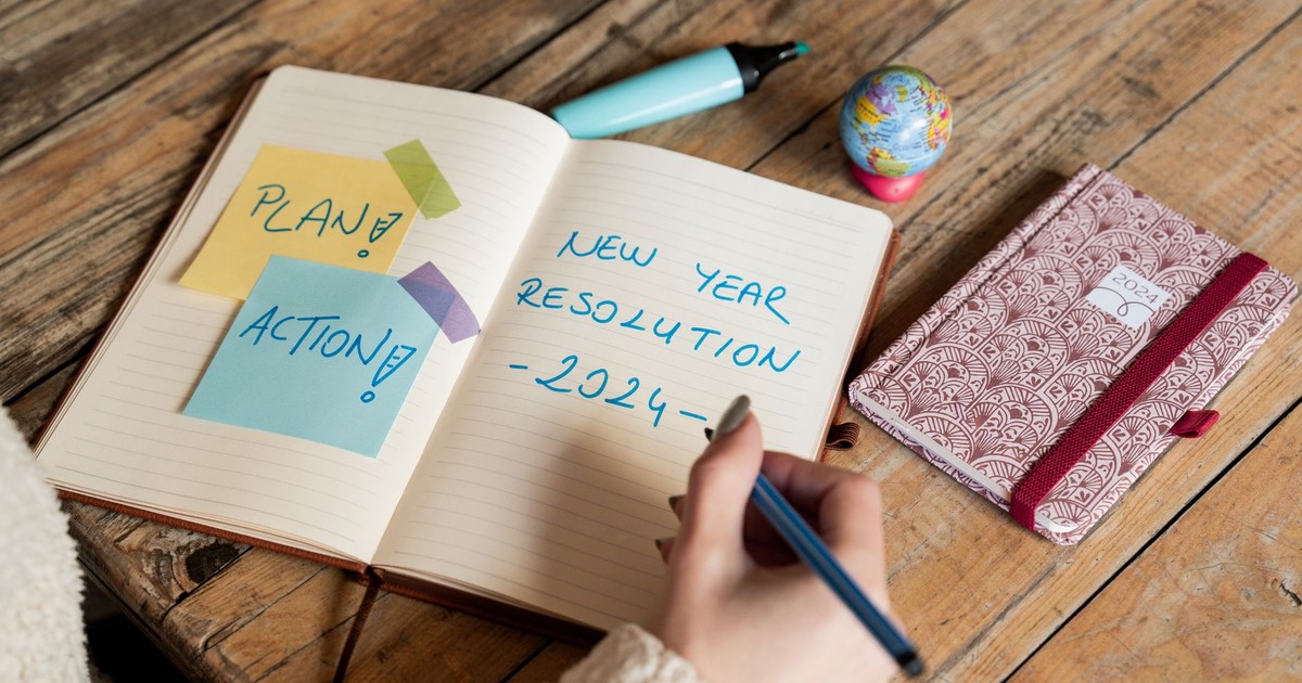 Person writing New Year 2024 resolutions in a notebook with sticky notes for planning and action.