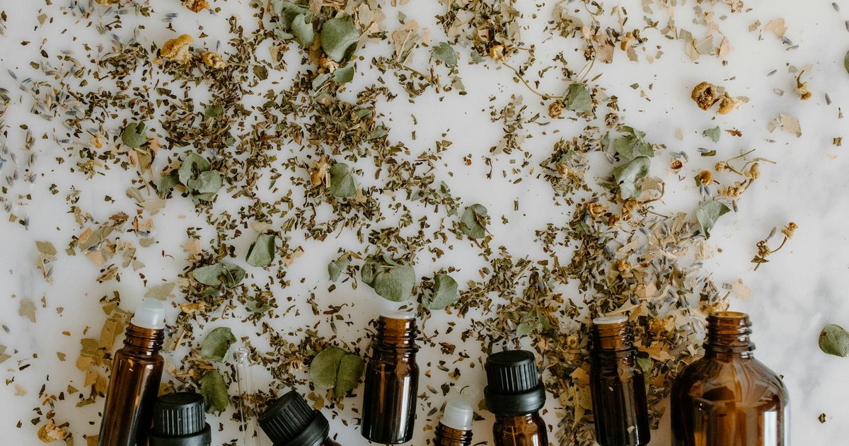 Glass bottles with essential oils surrounded by scattered dried herbs on a white surface.
