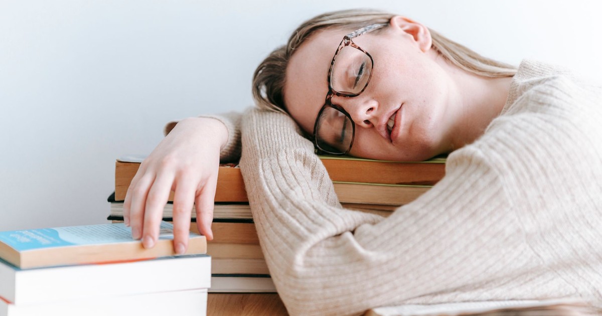 Exhausted female student in eyeglasses and casual sweater sitting at table and sleeping on books on light room