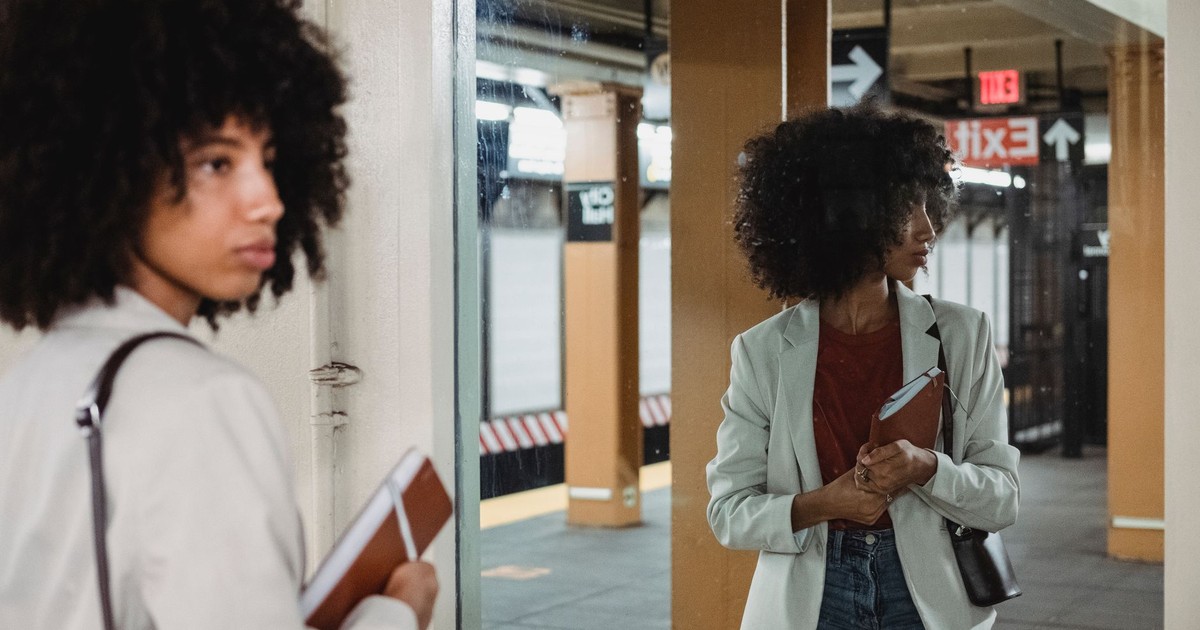 A fashionable woman with curly hair and a notebook reflected in a subway mirror.