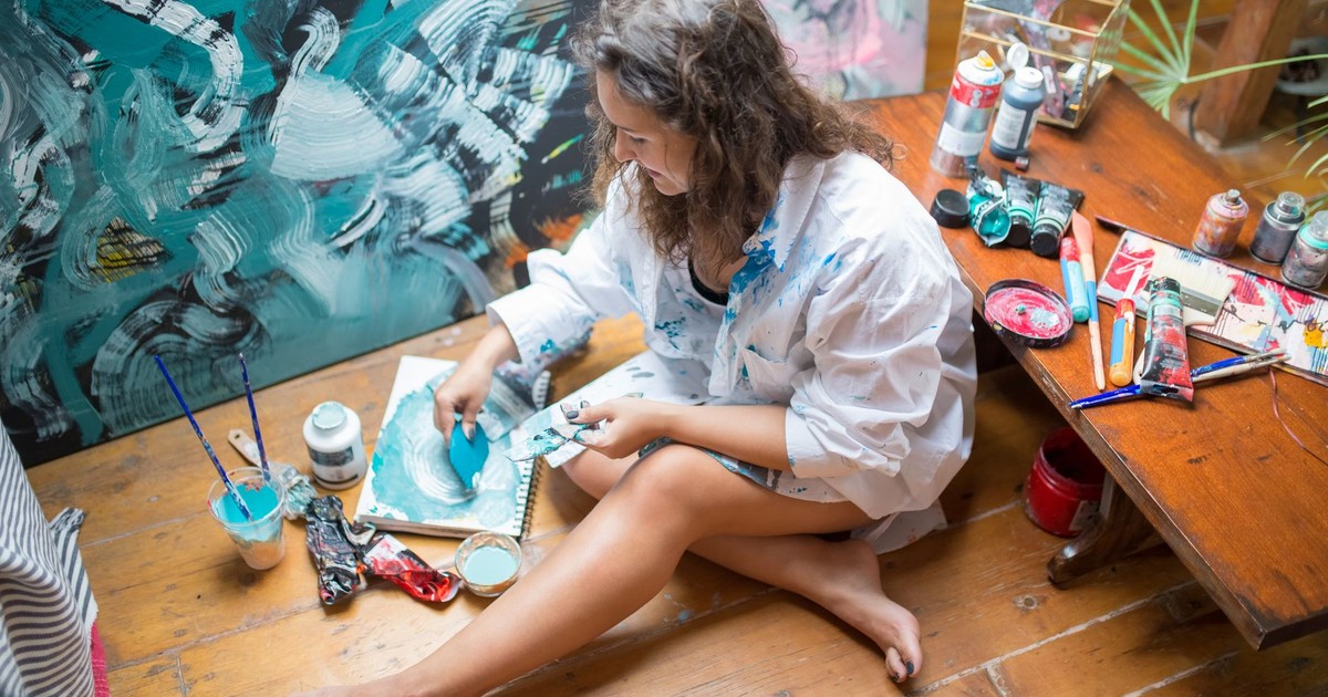 A woman artist painting abstract blue artwork indoors, surrounded by art supplies.