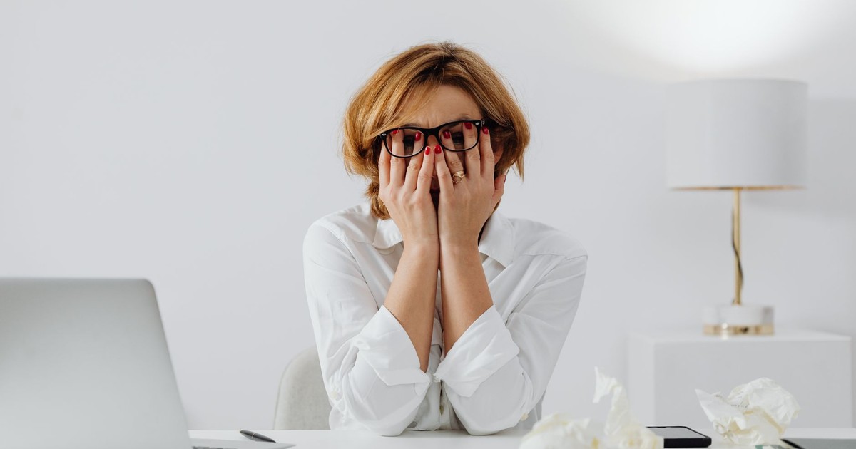 Woman with hands on face, sitting at desk with laptop and tissues, feeling stressed.