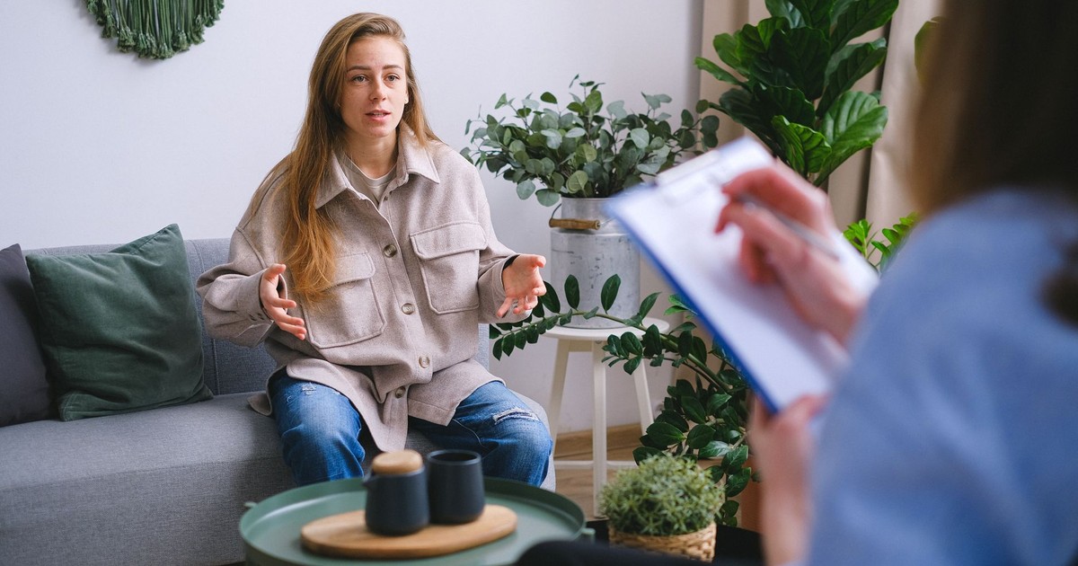 Concerned female client siting on couch and speaking about mental problems during psychotherapy appointment with blurred psychologist in light office