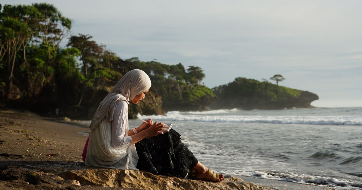 A woman in a hijab reads by the beach in Jawa Barat, Indonesia during a serene day.